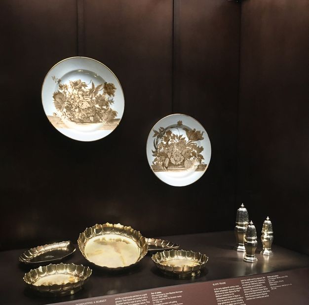 Installation. Two porcelain plates and eight pieces of metalwork are displayed in a case set into a wall. The plates hang on the wall over five gold-coloured dishes and three silver casters. Explanatory text can be seen (but not read) below the display.