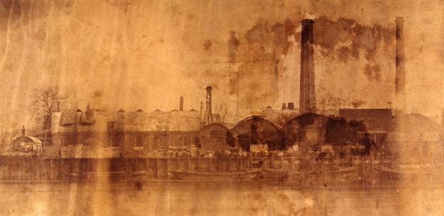 Photograph. This copper-toned photograph is streaked on its surface. A three-arched building and two smokestacks take up the right half of the image. More buildings are outlined against a blank sky to our left.