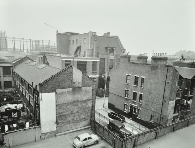Black and white photograph. We look down and across at several buildings situated close to each other with more structures beyond.