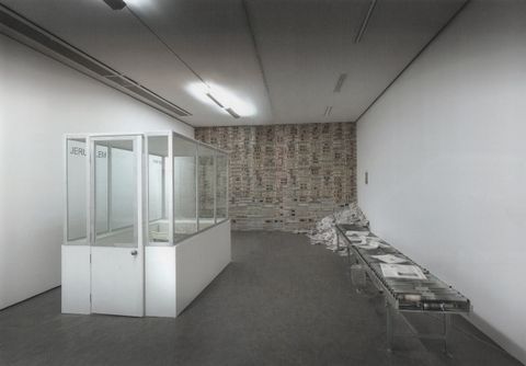 Installation. To our left, a glass booth with a white base faces a wall of newspapers at the end of the room. A few newspapers are laid out along a conveyor belt to our right and a pile of loose papers is at the end, in front of the wall of newspapers.