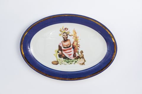 Porcelain. A dark-skinned woman wearing a low-cut pink dress smiles at the centre of this white oval plate. She is surrounded by plants and perhaps a seed. An illustration of a dark-skinned woman kneeling and holding hands together is to our right. The cobalt-blue rim is edged in gold.