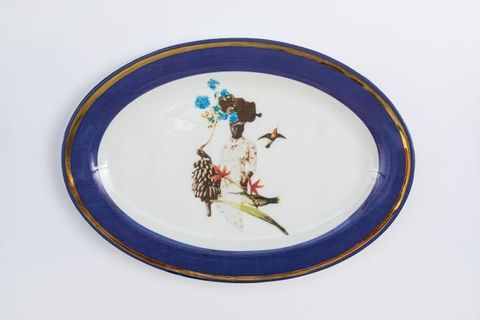 Porcelain. A dark-skinned woman is surrounded by two hummingbirds, a stalk of bananas, and two red flowers at the centre of this white oval plate. A basket of vivid blue flowers balances on her head. The cobalt-blue rim is edged in gold.