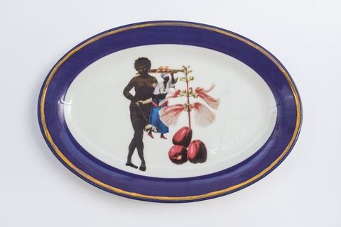 Porcelain. A nude, dark-skinned woman holds up a puppet or doll next to oversized flowers at the centre of this white oval plate. The cobalt-blue rim is edged in gold.