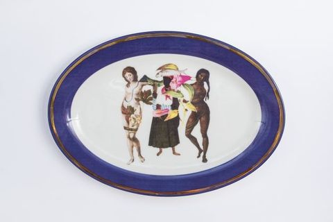 Porcelain. Two dark-skinned woman and a pale-skinned woman stand in a row at the centre of this white oval plate. One dark- and one pale-skinned woman are nude or nearly nude, and the other appears to be laden with fruit or flowers. The cobalt-blue rim is edged in gold.