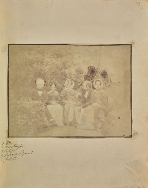 Black and white photograph. Two men and three women sit in a row facing us, in a garden or park setting. The image is pale shades of ash brown, mostly faded with a few notably dark areas.