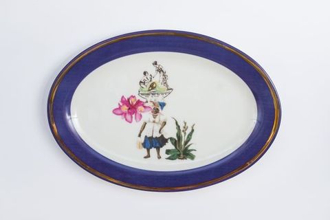 Porcelain. A dark-skinned woman supports an oversized fuchsia-pink flower and a tray that supports two figurines at the centre of this white oval plate. The figurines show on person kneeling and looking up at a woman who stands and bends towards the other. The cobalt-blue rim is edged in gold.