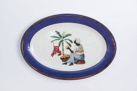 Porcelain. A dark-skinned woman sits near a palm tree holding yellow fruit in one hand at the centre of this white oval plate. She wears a white head covering and shirt, and blue-and-white striped, wide-legged pants. The cobalt-blue rim is edged in gold.