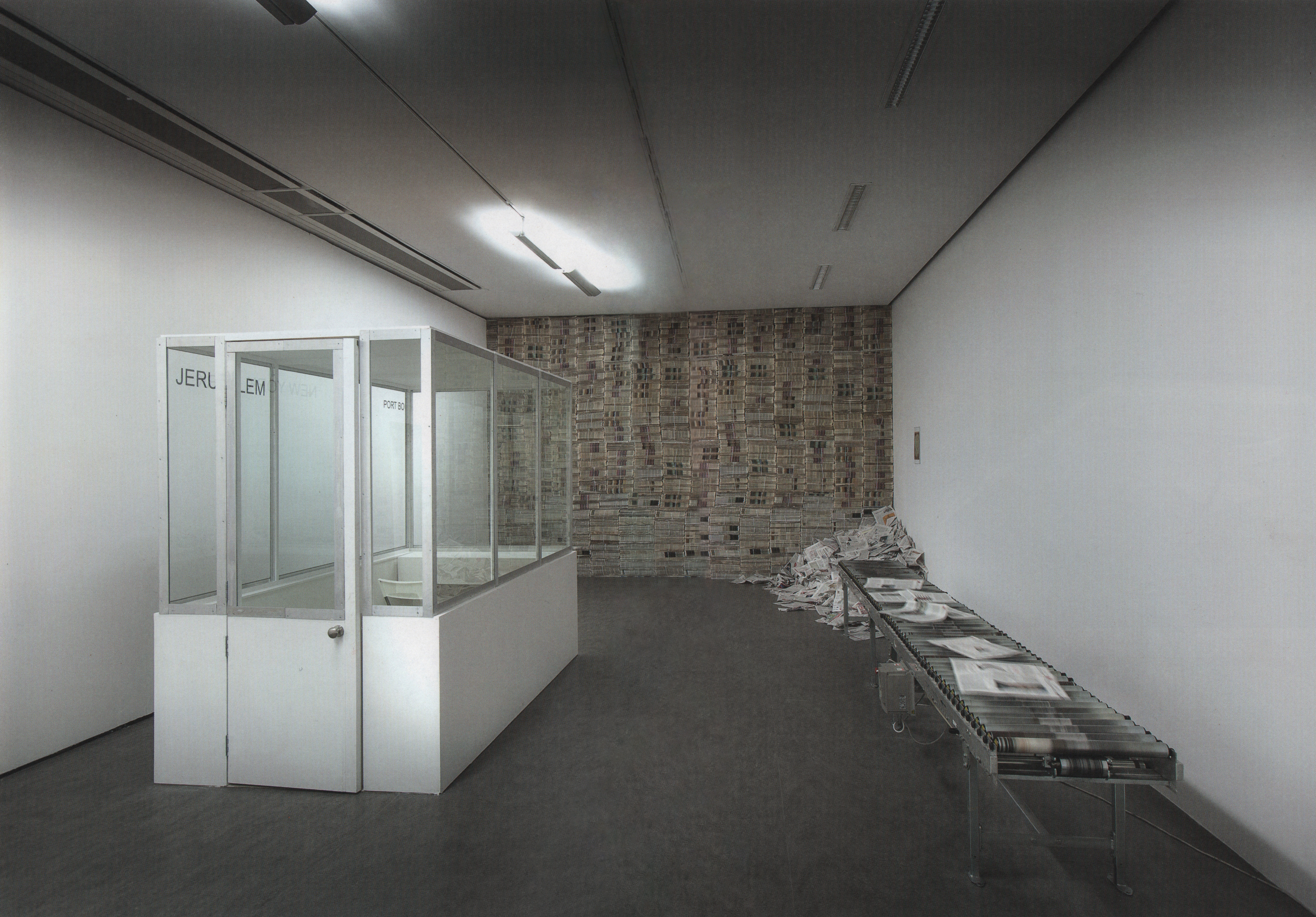 Installation. To our left, a glass booth with a white base faces a wall of newspapers at the end of the room. A few newspapers are laid out along a conveyor belt to our right and a pile of loose papers is at the end, in front of the wall of newspapers.