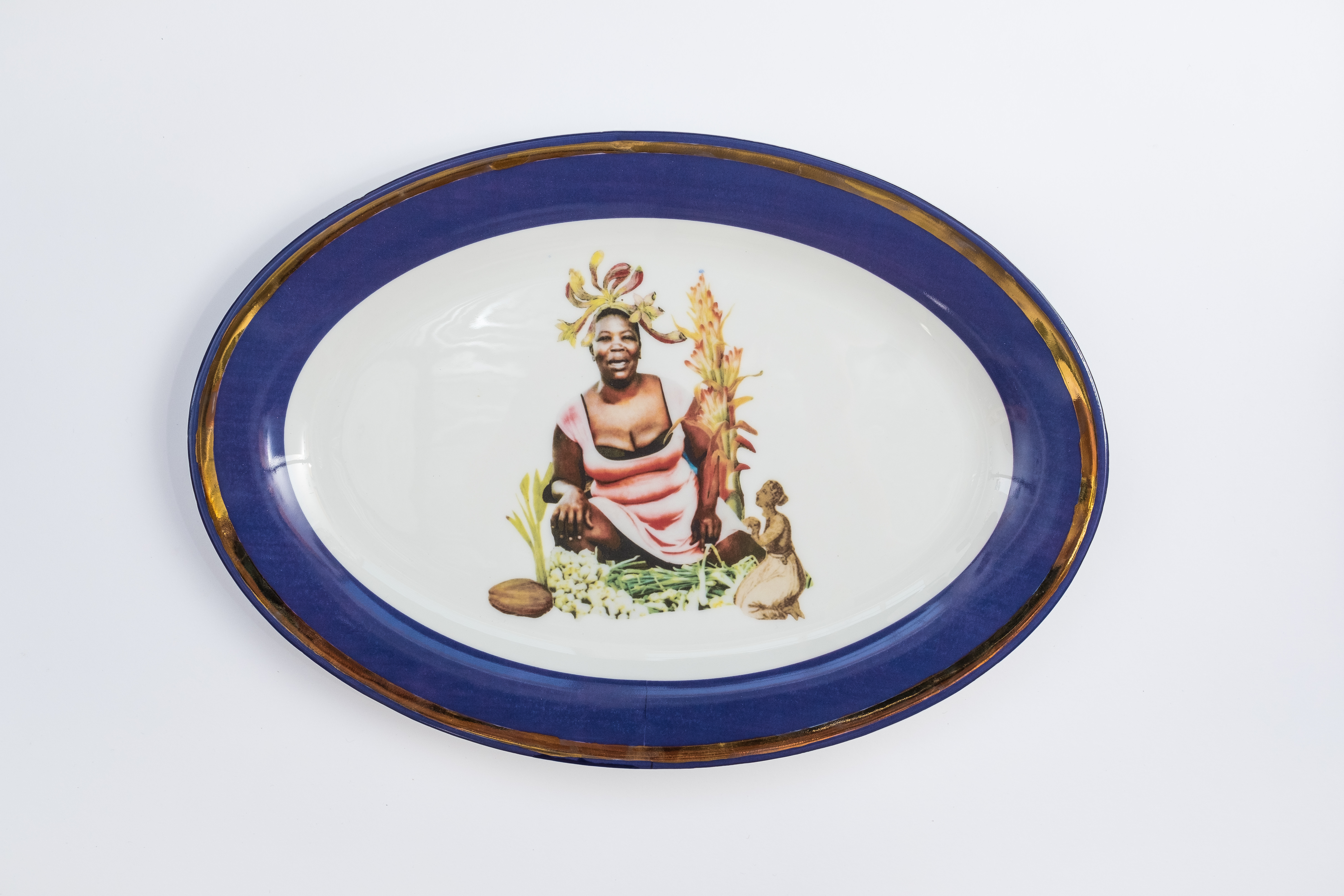 Porcelain. A dark-skinned woman wearing a low-cut pink dress smiles at the centre of this white oval plate. She is surrounded by plants and perhaps a seed. An illustration of a dark-skinned woman kneeling and holding hands together is to our right. The cobalt-blue rim is edged in gold.