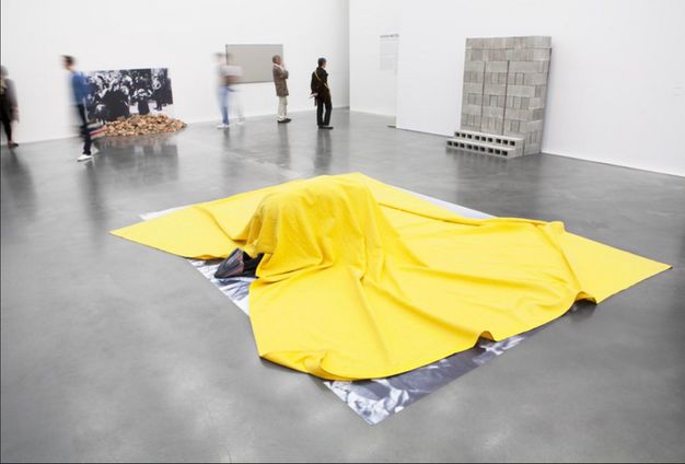 Installation. An enlarged black and white photograph is laid on the grey floor of a gallery. A tarp-sized sheet of buttercup-yellow fabric lies over the photograph. Here, a person has crawled under the cloth and only the bottoms of their sneakers stick out.