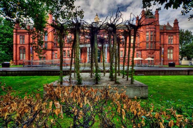 Sculpture. Five rows of slender tree trunks are set into a slab of concrete so their roots thrash against the sky. The concrete slab sits on a grassy lawn. A stately red brick building in the background nearly spans the width of the photograph.
