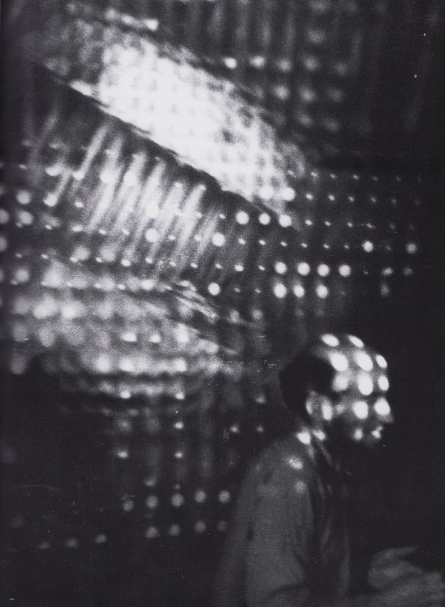 Black and white photograph. A person may be lit by holes in perforated material that also shine on the surface behind him, or may stand in front of perforated material in this vertical photograph.