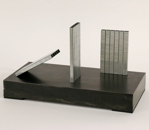 Sculpture. Two panels of staples stand on a black base at right angles to each other. A third angles at about thirty degrees, leaning towards the others.