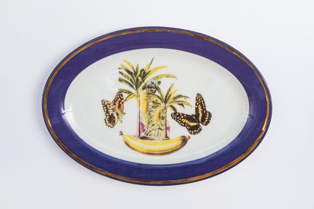 Porcelain. A dark-skinned person is nearly hidden behind patterns spanning two palm trees that stand on an out-scaled banana at the centre of this white oval plate. Two black and yellow butterflies flit to each side. The cobalt-blue rim is edged in gold.