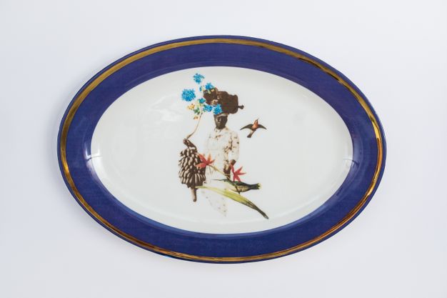 Porcelain. A dark-skinned woman is surrounded by two hummingbirds, a stalk of bananas, and two red flowers at the centre of this white oval plate. A basket of vivid blue flowers balances on her head. The cobalt-blue rim is edged in gold.