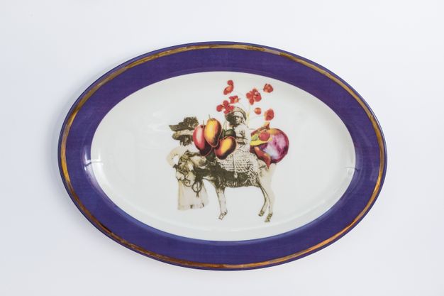 Porcelain. A dark-skinned woman sits on a donkey and the other stands by its head. They are surrounded by red and yellow fruits, vegetables, or flowers at the centre of this white oval plate. The cobalt-blue rim is edged in gold.