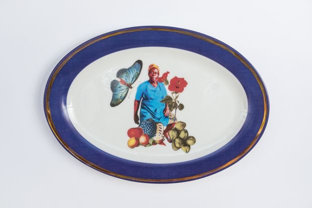 Porcelain. A dark-skinned woman wearing blue stands at the centre of this white oval plate. She is surrounded by a sprig of red flowers, a blue butterfly, and perhaps fruit and seeds. The cobalt-blue rim is edged with gold.