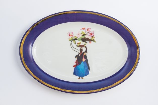 Porcelain. A dark-skinned woman holding flowers balanced on her head wears a black shawl over a flaring blue skirt at the centre of this white oval plate. The cobalt-blue rim is edged in gold.