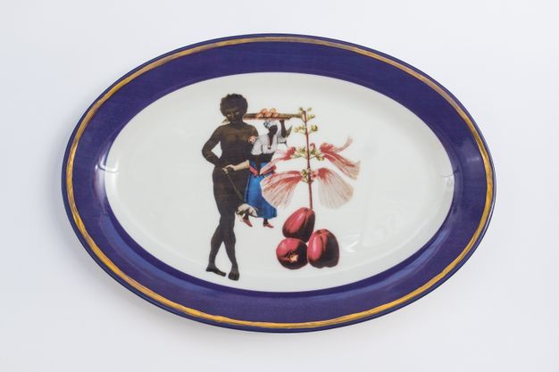 Porcelain. A nude, dark-skinned woman holds up a puppet or doll next to oversized flowers at the centre of this white oval plate. The cobalt-blue rim is edged in gold.