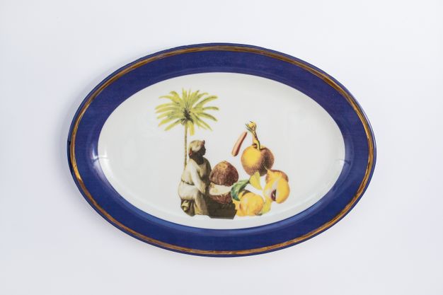 Porcelain. A dark-skinned woman wearing white sits with an oversized coconut and fruit under a palm tree at the centre of this white oval plate. The cobalt-blue rim is edged in gold.