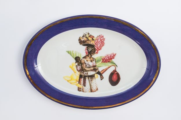 Porcelain. A woman carries a baby on her back at the centre of this white oval plate. Both are dark skinned. The woman balances a basket of fruit on her head and is backed with an oversized flower. The cobalt-blue rim is edged in gold.