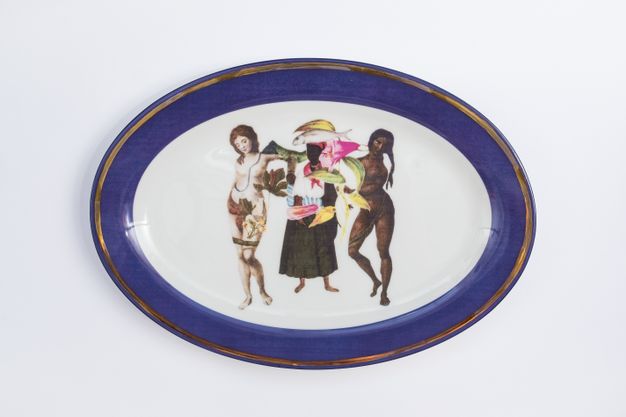 Porcelain. Two dark-skinned woman and a pale-skinned woman stand in a row at the centre of this white oval plate. One dark- and one pale-skinned woman are nude or nearly nude, and the other appears to be laden with fruit or flowers. The cobalt-blue rim is edged in gold.