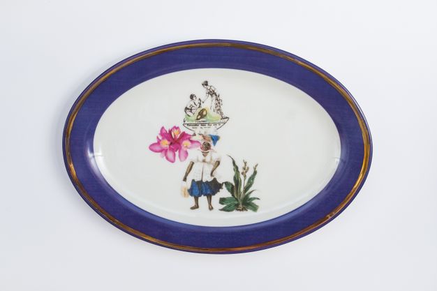 Porcelain. A dark-skinned woman supports an oversized fuchsia-pink flower and a tray that supports two figurines at the centre of this white oval plate. The figurines show on person kneeling and looking up at a woman who stands and bends towards the other. The cobalt-blue rim is edged in gold.
