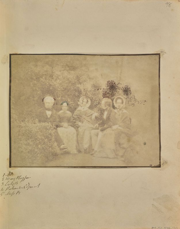 Black and white photograph. Two men and three women sit in a row facing us, in a garden or park setting. The image is pale shades of ash brown, mostly faded with a few notably dark areas.