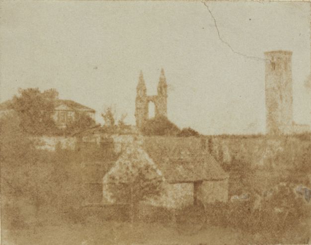 Black and white photograph. In tones of bronze and fawn brown, a small structure sits in front of a wall the same height as its pitched roof. A tower and ruins of a cathedral are outlined against the blank sky in the town beyond the wall.