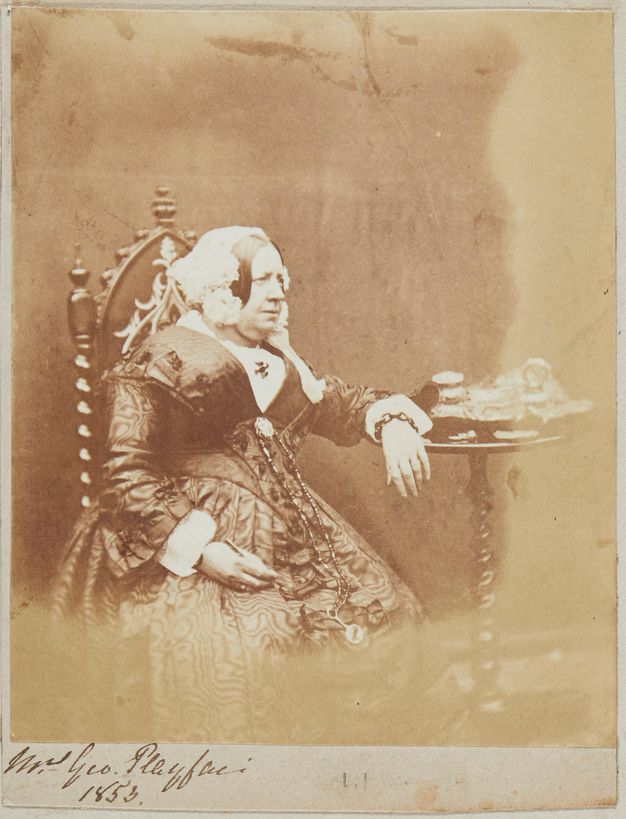 Black and white photograph. A woman wearing a white bonnet looks off to our left, her lips slightly parted over a double chin. One arm rests on a round table next to her, and the other is in her lap, perhaps holding a pen.