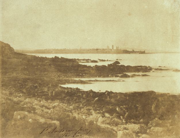 Black and white photograph. A rocky shoreline zigzags from the lower right corner back to the centre of this photograph. The water appears smooth and white. A low stretch of land with a few buildings is along the horizon.