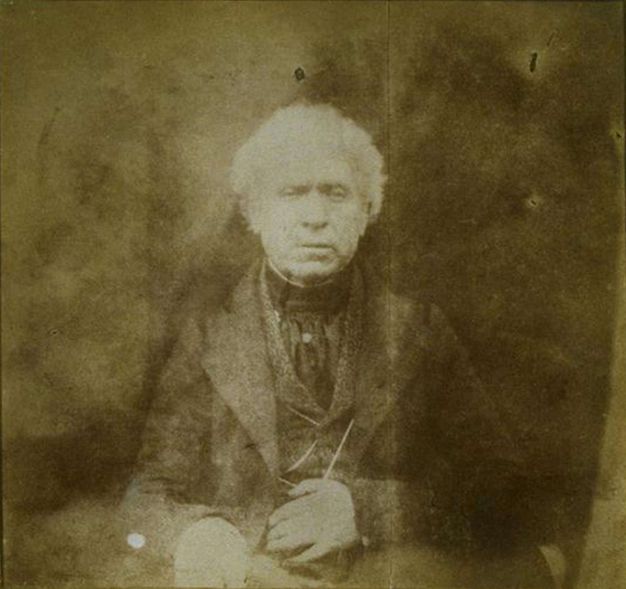 Black and white photograph. Shown from the lap up, a pale-skinned, clean-shaven man with white hair wears a three-piece suit. He rests one hand on his belly, the other on his knee. He looks slightly down with lips parted.