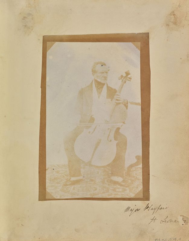 Black and white photograph. In tones of copper brown and tan, a man sits behind a cello, the bow resting on the strings. The other hand is on the neck of the instrument where it meets the body. The pale-skinned, clean-shaven man looks off to our right.