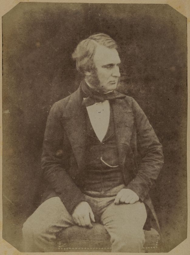 Black and white photograph. A pale-skinned man with mutton chops sits and looks off to our right. One hand rests at the top of his thigh, the other on the upholstered seat between his legs.