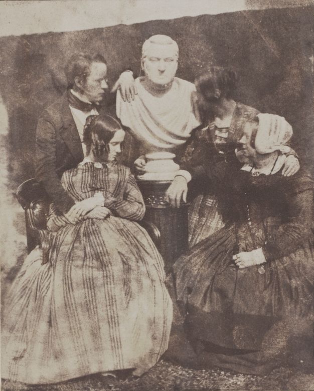 Black and white photograph. Three women and a man, all pale-skinned, gather around a marble bust of another man. One woman wearing a white bonnet rests a forearm on the pedestal base of the bust, and a younger woman wraps one hand around the backside of the base. The living man drapes one arm over the bust’s shoulder and rests the other on a young woman’s lap, who sits in front of him.