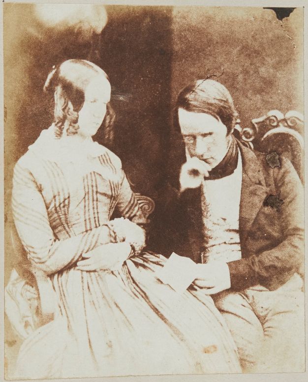 Black and white photograph. Shown from the laps up, a pale-skinned man and woman, both with dark hair, sit together. The man rests his chin in one hand, pince-nez balanced on the bridge of his nose as he reads a letter. The woman crosses her wrists, leans back in her seat, and gazes off to our right.