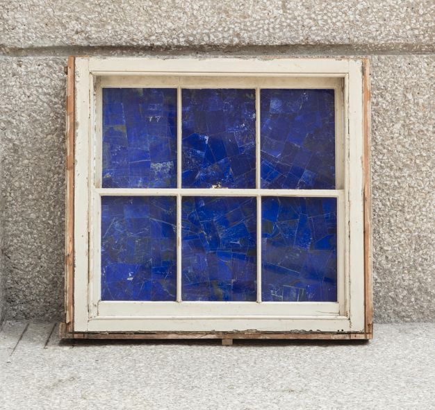 Sculpture. The openings of a six-paned window painted off-white are filled with pieces of lapis lazuli, so they practically glow blue. The window leans up against a stone or concrete wall.