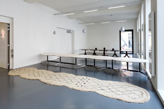 Installation. A long beige-coloured rug with uneven edges lies before two rows of tables that each have one continuous tabletop supported by sleek metal or wooden legs.