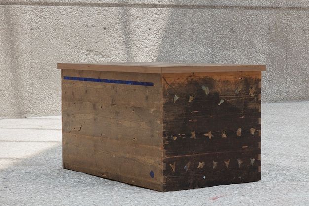 Sculpture. A wooden box is made with horizontal planks of dark wood. A strip of vivid blue is near the flat lid on the long side to our left. Objects like bits of twigs or shells may be affixed to the short side to our right, but details are difficult to make out.