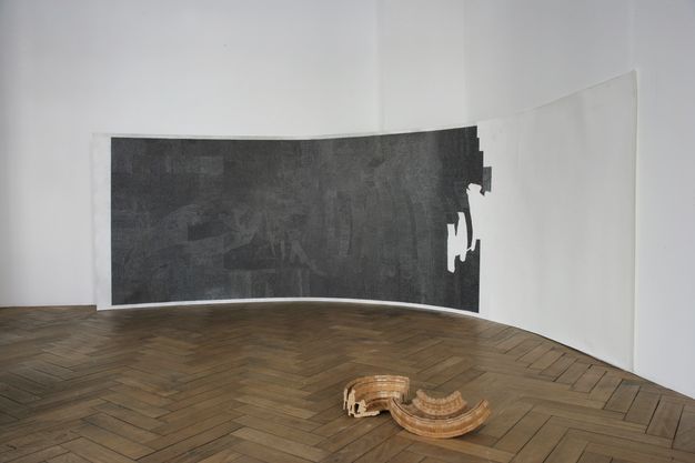 Installation. A long piece of white paper rests on the wood flfjbishopoor of a gallery and curves along a corner of the room. The left two-thirds is coloured black; the right third is white. Two pieces of a ceiling medallion lie on the floor closer to us.