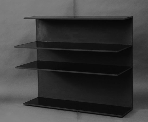 Black and white photograph. Four shelves float in front of a backing board, like a bookcase without sides. The object is angled to our left so those corners of the shelves stick out like triangular blades of an exacto knife.