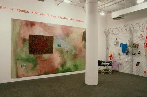 Installation. A mural painting hangs on a wall beneath red text. A section of chain-link fencing topped with coiling barbed wire is to our right. A baby pram sits in front of the fence, which is hung with colourful objects.