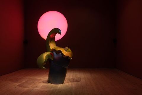 Installation. A sculpture has smooth surfaces in midnight blue and golden sage. A columnar base supports a ruffled line, wing-like projection, and a curving hook. A carnation-pink disc glows behind it. The gallery is otherwise dim except for spotlights shining down on the sculpture.