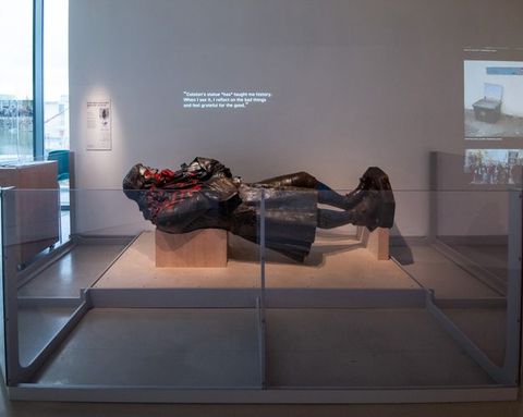 Installation. A bronze sculpture of a standing man is displayed lying across wooden supports in a gallery. The statue has been spray painted with red on the face and across the chest. Text projected onto the wall above it reads, 'Colston’s statue *has* taught me history. When I see it, I reflect on the bad things and feel grateful for the good'.