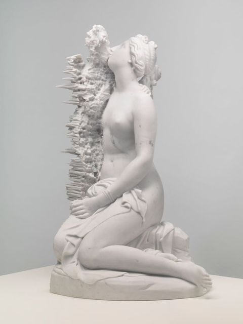 Sculpture. A mostly nude woman kneels with a cloth across her hips. Her hair is plaited, and her face is lifted, facing our left in profile. A form next to her is difficult to make out. It looks like vegetation along her body but turns into long spikes on the far side.