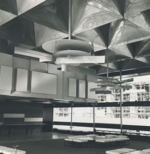 Architecture. Black and white photograph of an interior building filled with abstract designs, from the reflective three-dimensional grid ceiling, to the layered rectangular wall panels, and hanging mobile sculptures of circular plates and rectangular beams.