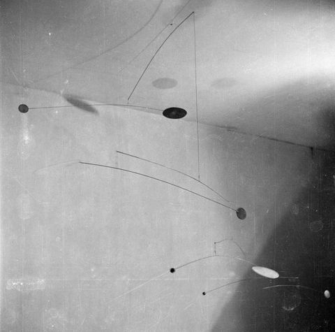 Metal sculpture. Black and white photograph looking up at a mobile sculpture hanging from the ceiling with multiple tiers of narrow curved metal rods and cables that offer a delicate and bouncy three-dimensional shape.