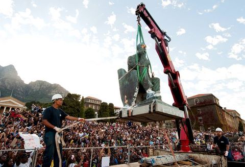 A crane picks up statue of a seated man as a worker wearing a hard hat guides it with a strap. Hundreds of people look on from the background under a blue sky.