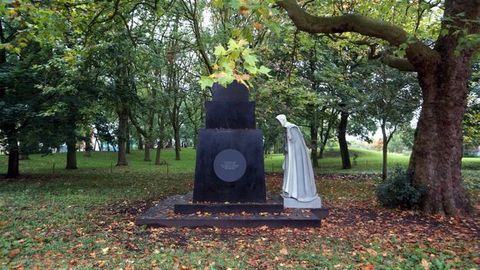 Public art. An empty black plinth is faced with a round plaque. A sculpted person wearing a long robe and white head covering bends to examine something on the plinth. The work is set up under a sprawling tree in a park.