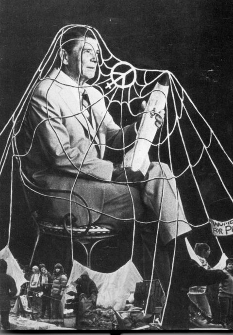 Photomontage. A spider web drapes over a man, Ronald Reagan, sitting with his legs crossed. He holds a missile in one hand. Out of scale, people stand near a tent and hold up protest signs around his feet.